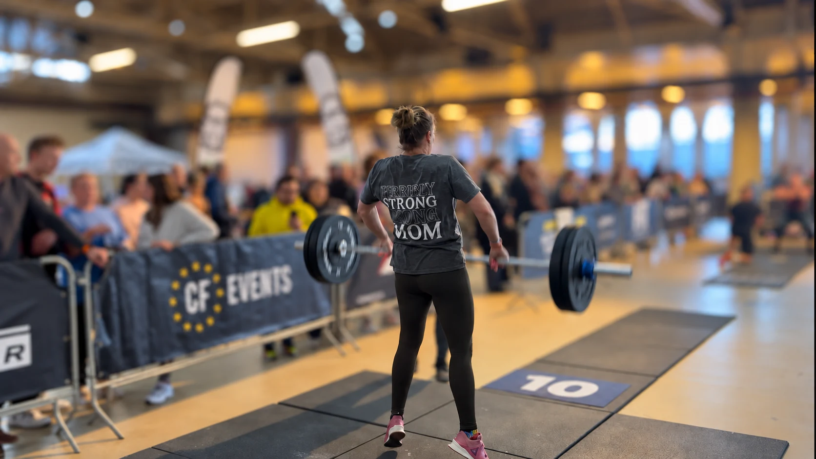 Een atleet tilt een halter tijdens een wedstrijd, met supporters langs de vloer.